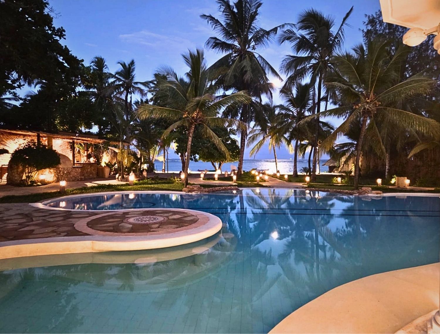 Poolside at a tropical resort at dusk, with palm trees, warm lights, and the ocean visible in the background. - Soroi Collection - Soroi Collection
