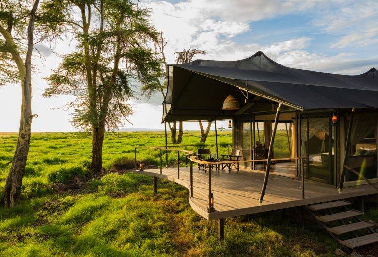 Tented lodge on a wooden deck in a grassy savanna, with trees in the background and outdoor seating under a dark canopy.