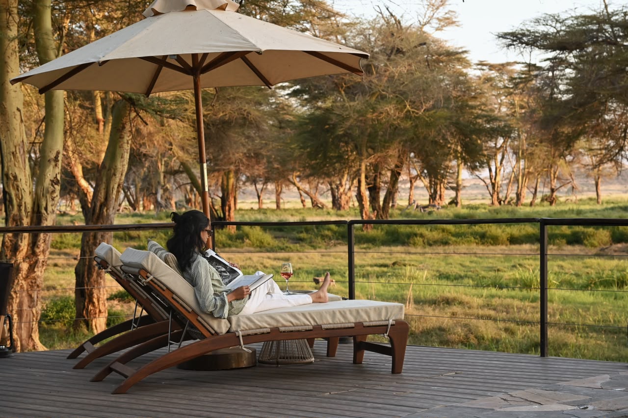 Person lounging on a cushioned outdoor lounge chair under a large umbrella on a wooden deck, reading a magazine with a glass of wine nearby. - Soroi Collection - Soroi Collection