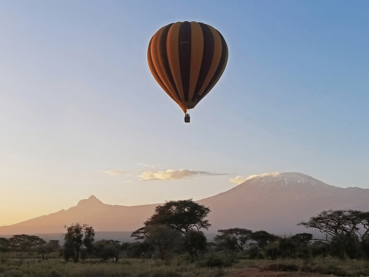 Striped orange and black hot air balloon drifting over a savannah with distant snow-capped mountain at sunset. - Soroi Collection
