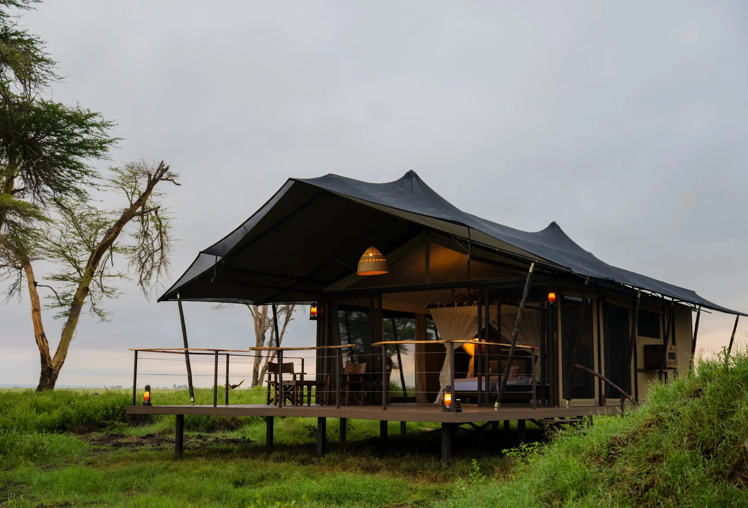 Raised wooden-deck pavilion with a black fabric roof, warm lanterns, and outdoor seating in a windy, grassy field with trees nearby, under an overcast sky. - Soroi Collection - Soroi Collection