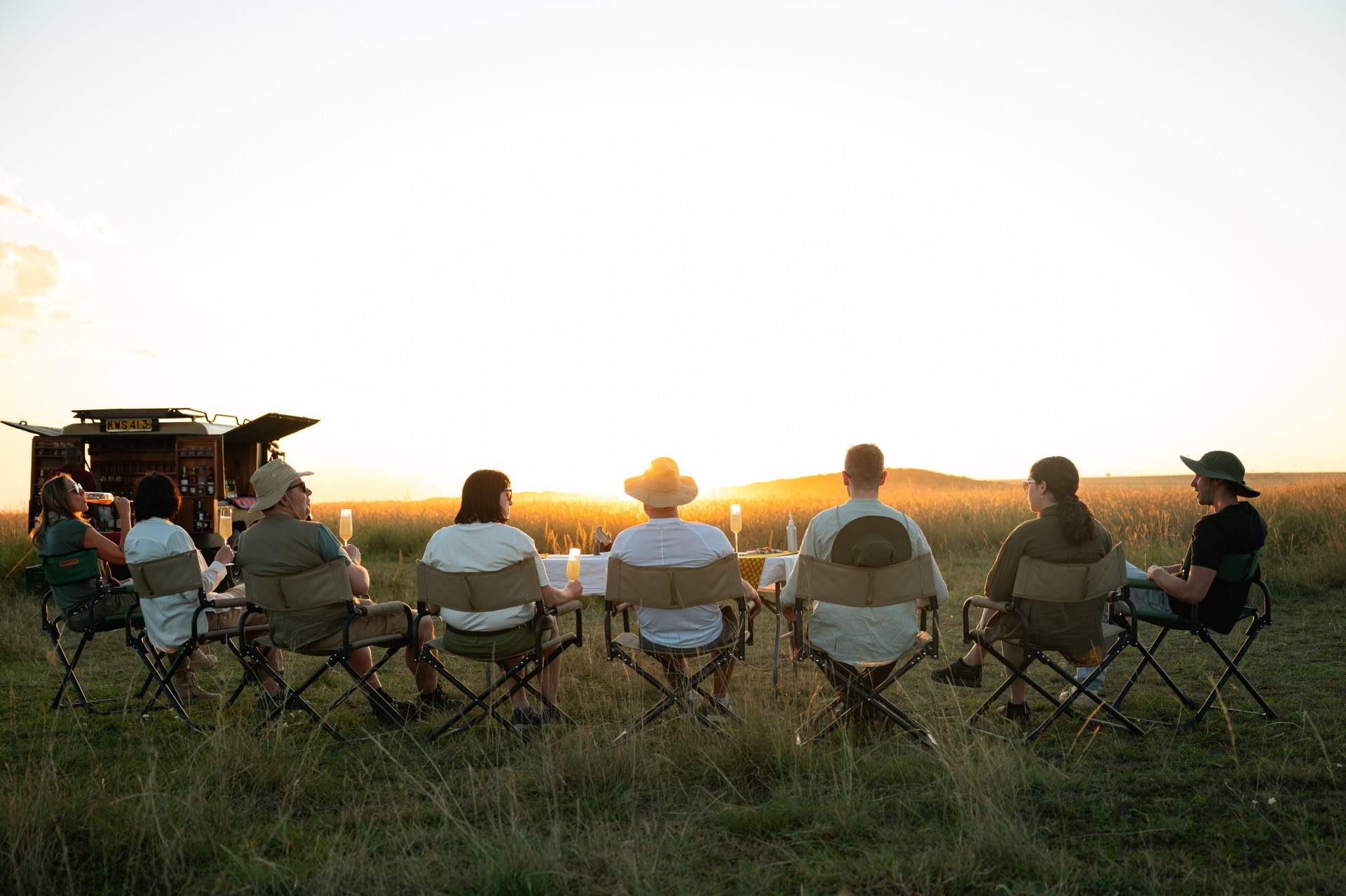 Group of people sitting in folding chairs around a long table in a grassy field at sunset, drinks and candles on the table. - Soroi Collection